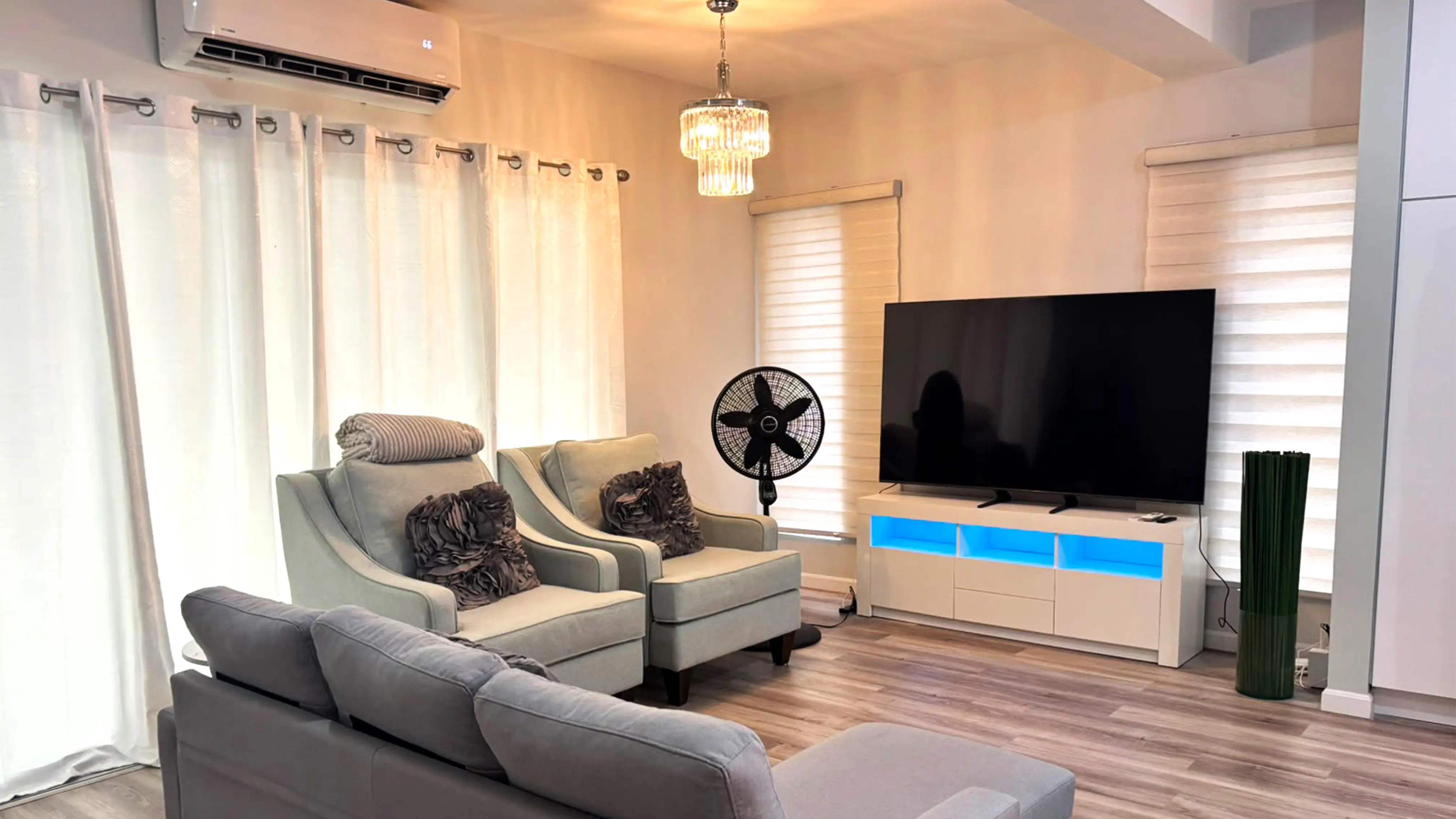 Furnished living room with a grey chaise sofa, two armchairs, television, chandelier and wall-mounted air conditioner.