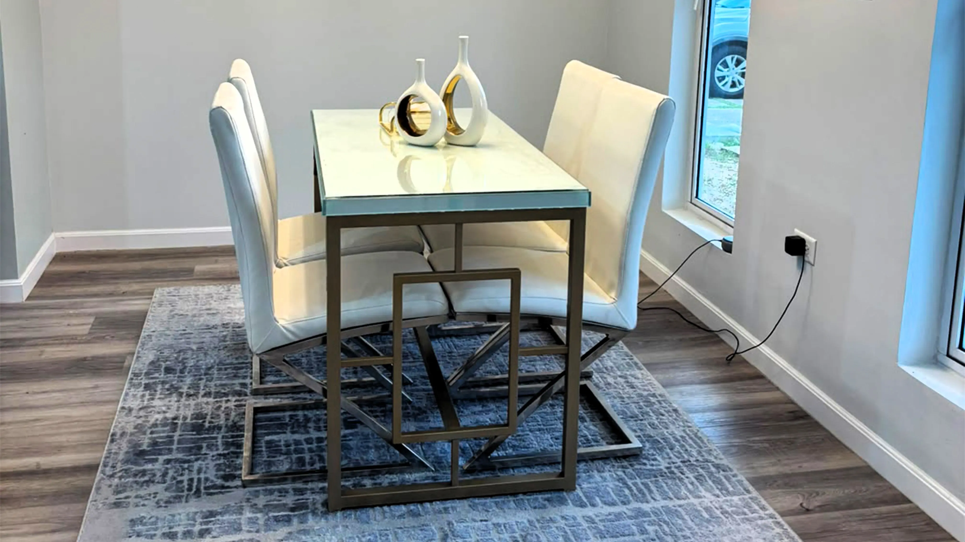 Dining table with four white upholstered chairs and decorative vases on a grey patterned rug beside tall windows.