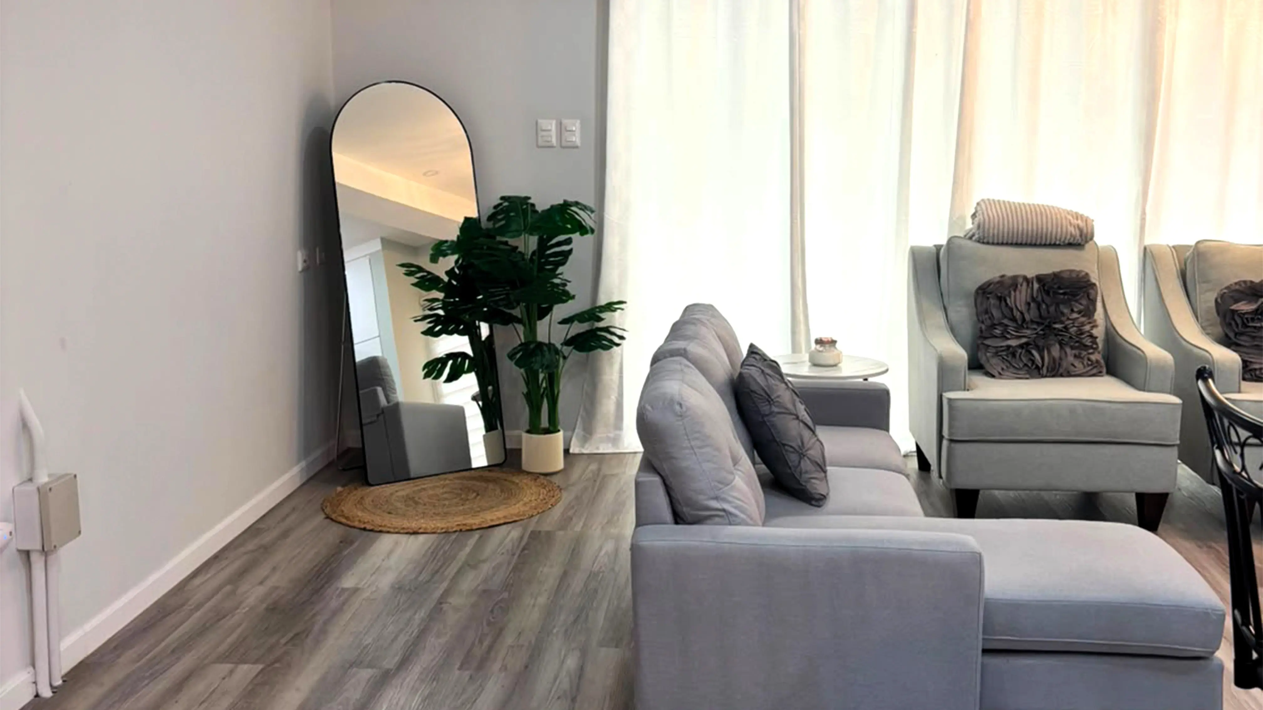 Grey chaise sofa and armchairs beside white curtains, with an arched floor mirror and potted plant in the corner.