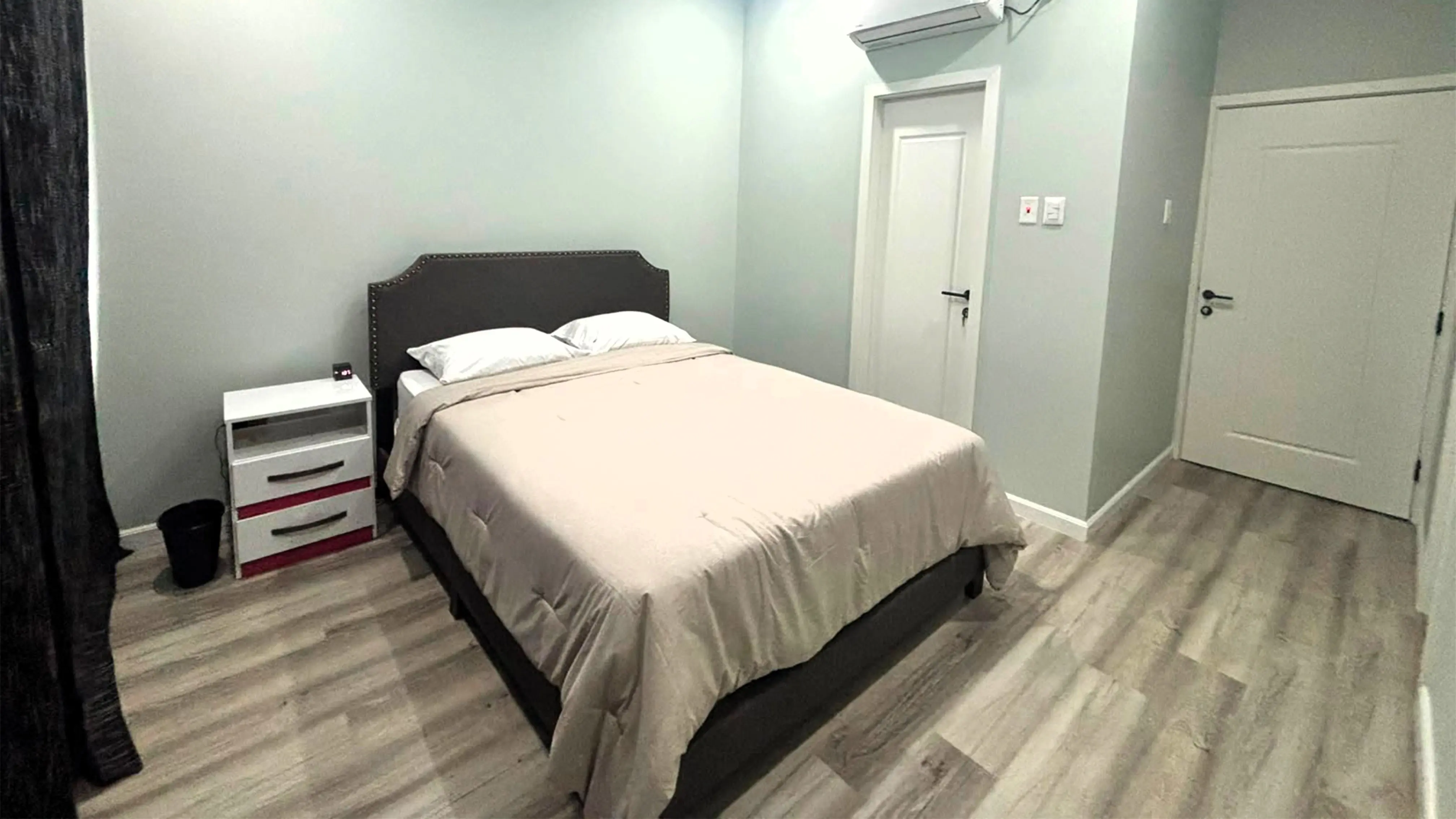 Bedroom with beige bedding, a dark upholstered headboard, bedside cabinet, wood-look flooring and wall-mounted air conditioner.