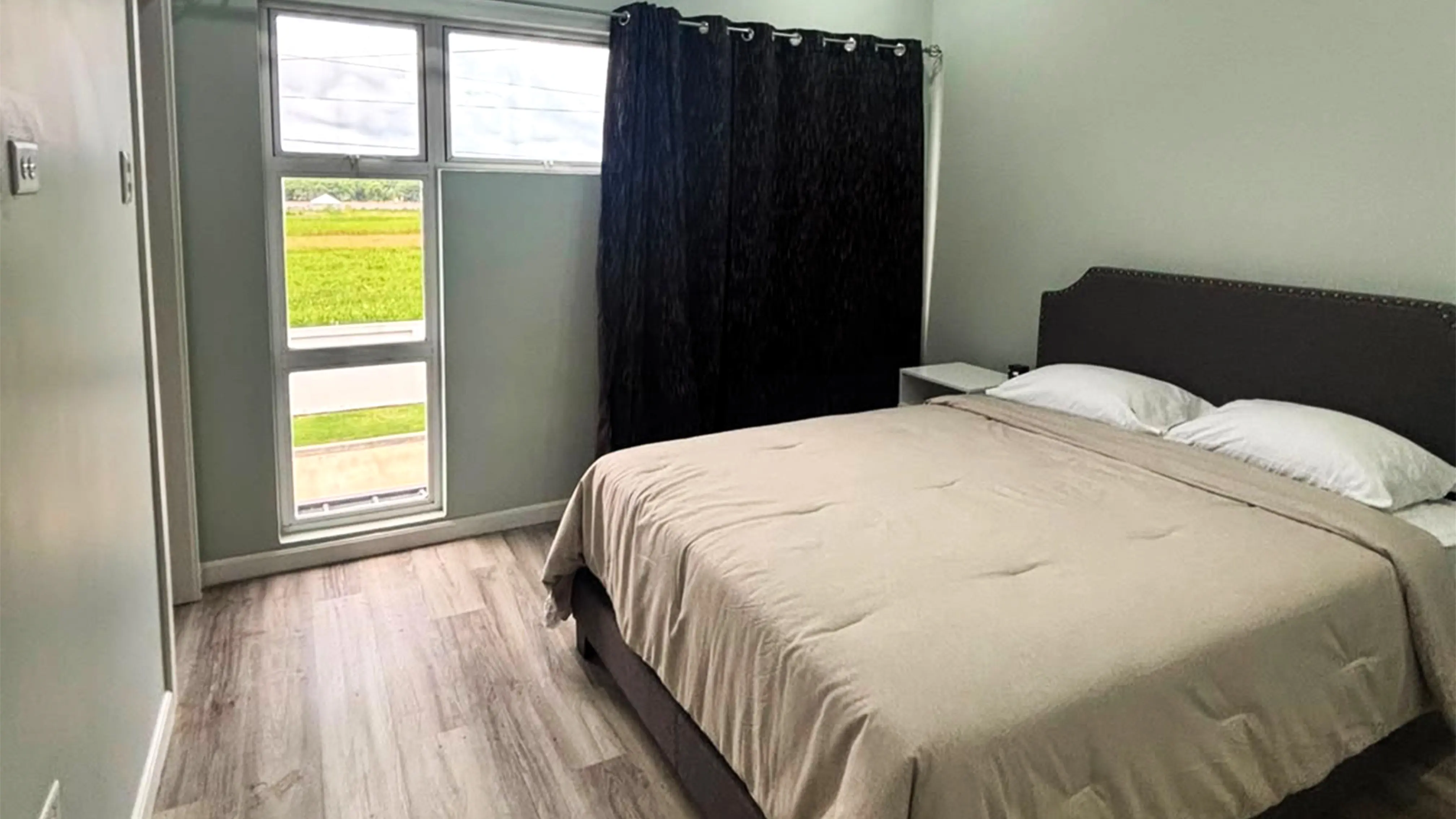 Bed with beige bedding beside tall windows and partly drawn dark curtains, with green fields visible outside.