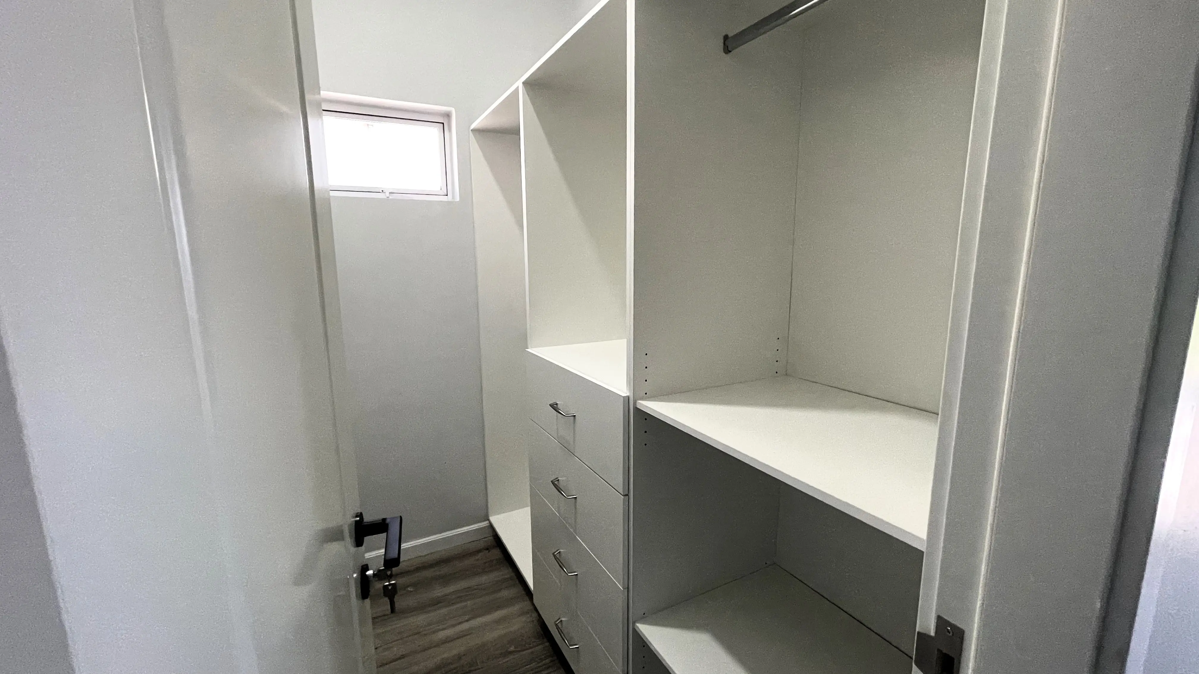 Closet with white built-in drawers, open shelves, hanging rails and a small high window.