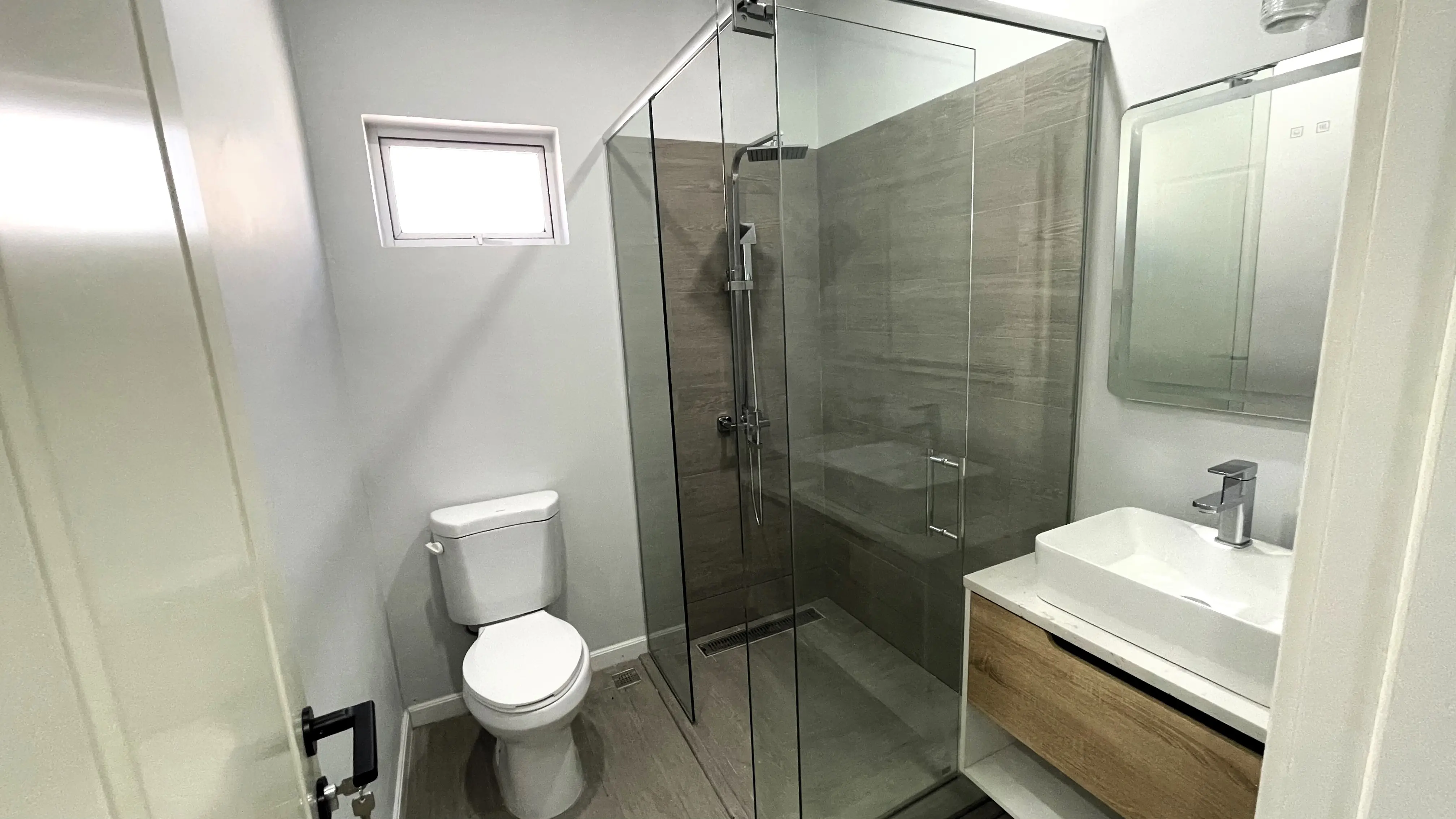 Bathroom with a glass-enclosed rainfall shower, grey wall tiles, white toilet and vessel sink on a wood-finish vanity.