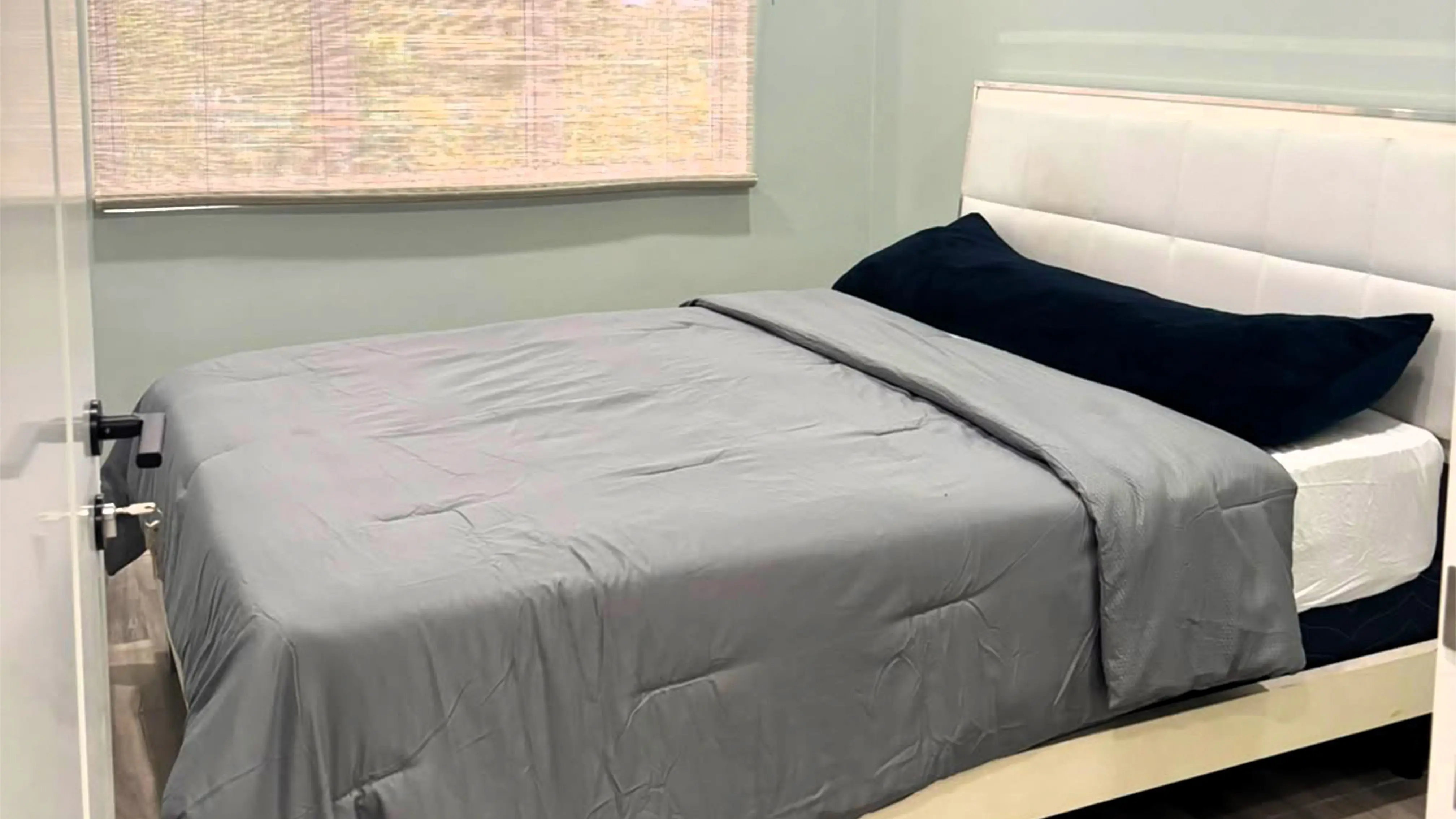 Bed with a white padded headboard, grey bedding and a navy pillow beside a window with a woven blind.