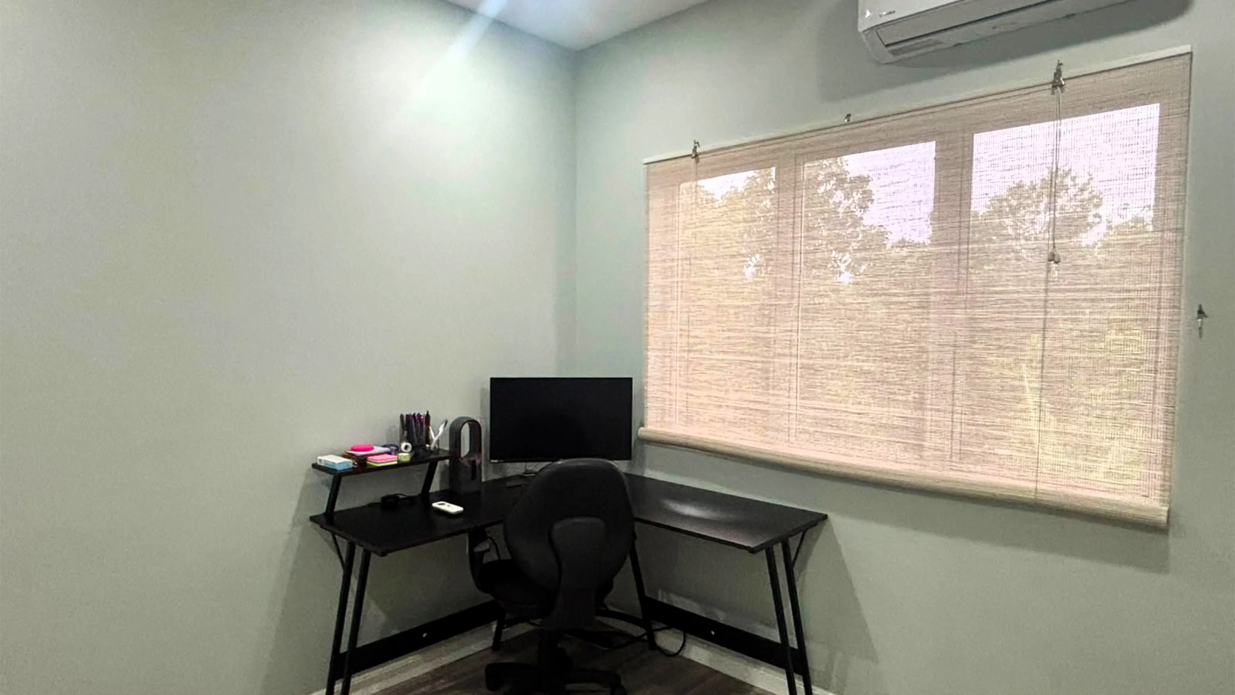 Home office with a black corner desk, monitor and swivel chair beneath a wide window and wall-mounted air conditioner.