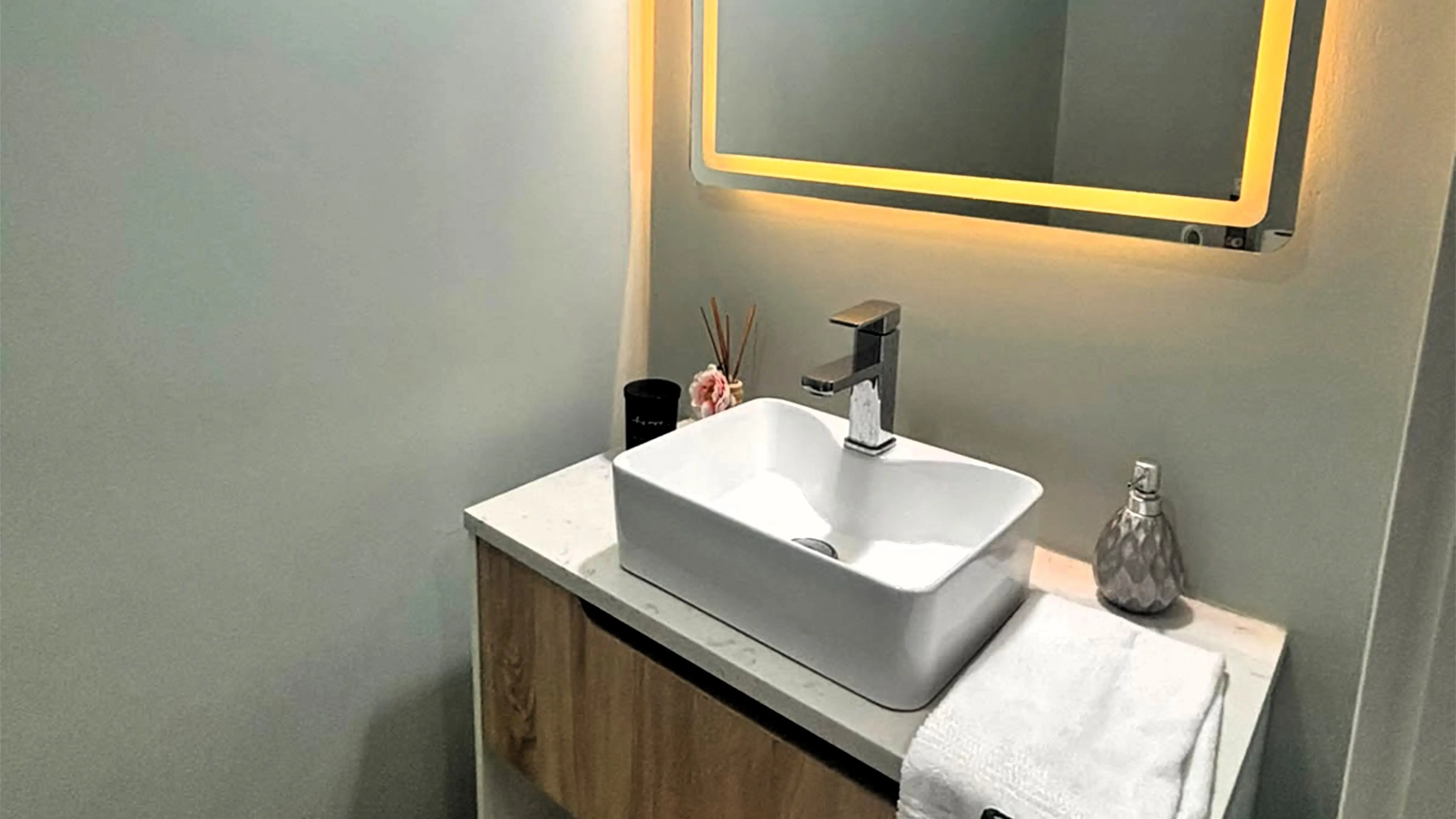 White vessel sink and chrome tap on a wood-finish vanity beneath a mirror with warm illuminated edging.
