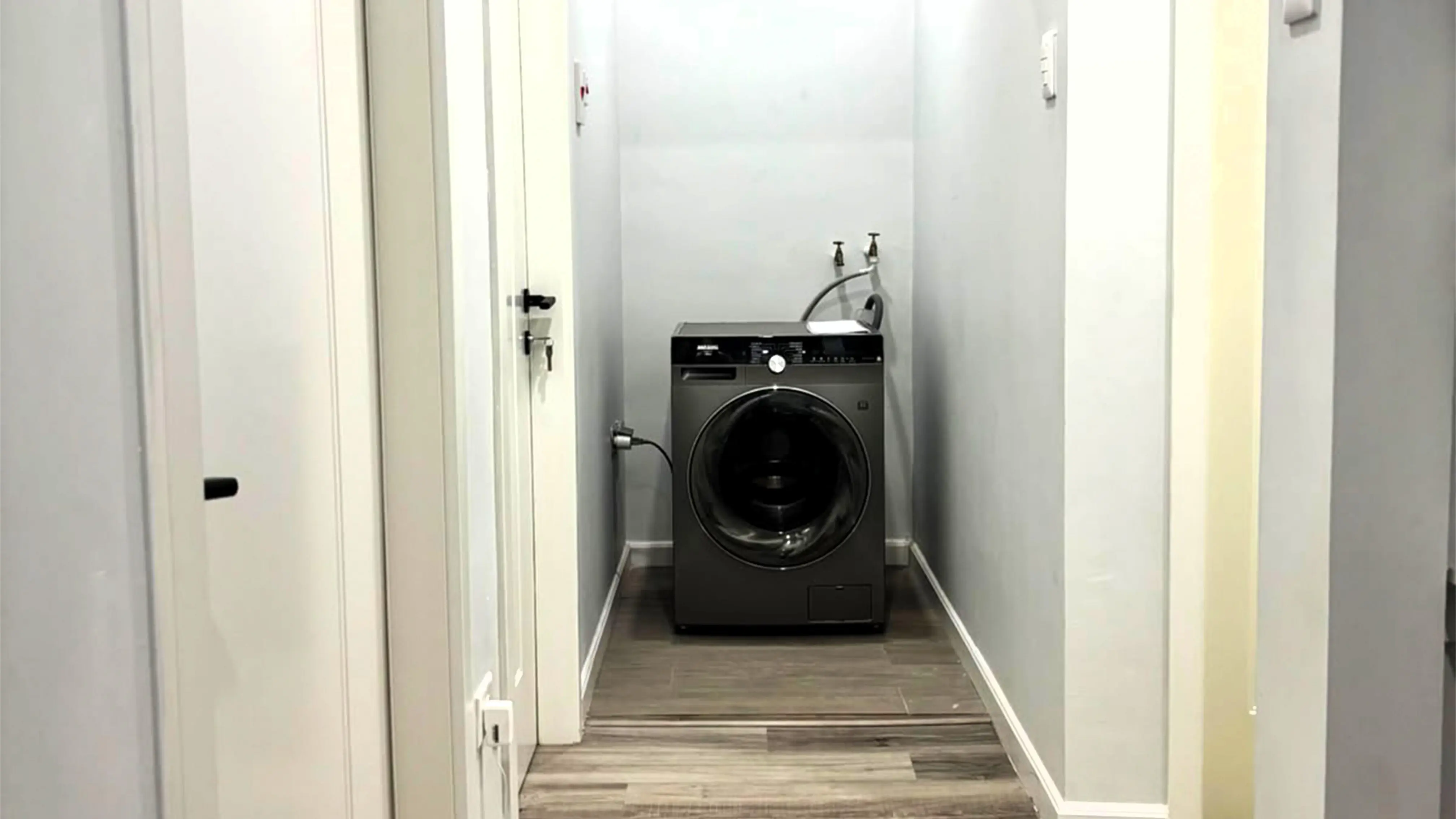 Laundry alcove at the end of a hallway with a dark front-loading washing machine and water connections above.