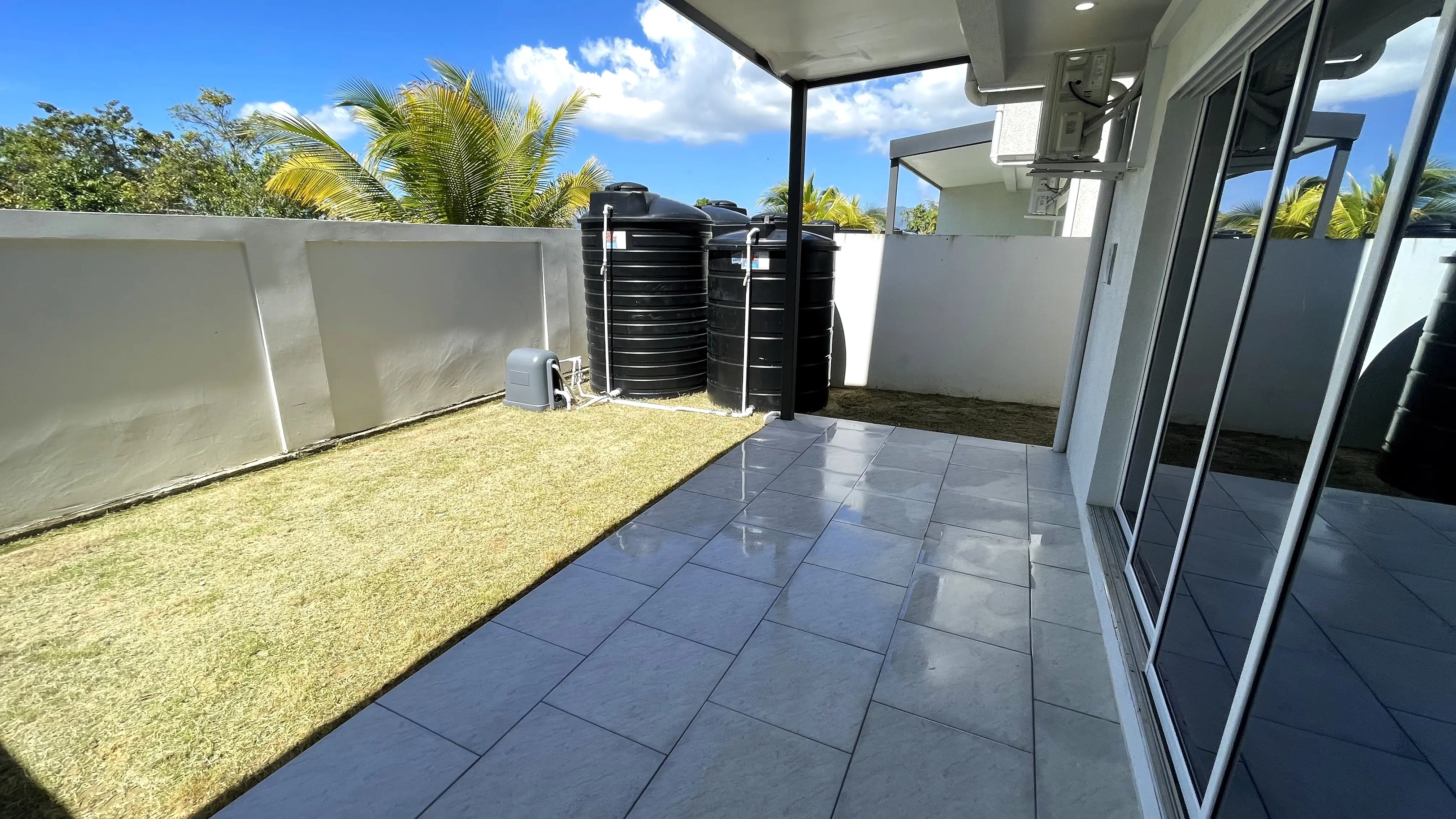 Enclosed yard with a grass strip, covered tiled patio and black water storage tanks beside the boundary wall.
